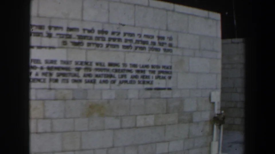 1960s Israel home-movie frame showing a pale stone wall with Hebrew and English inscription text.