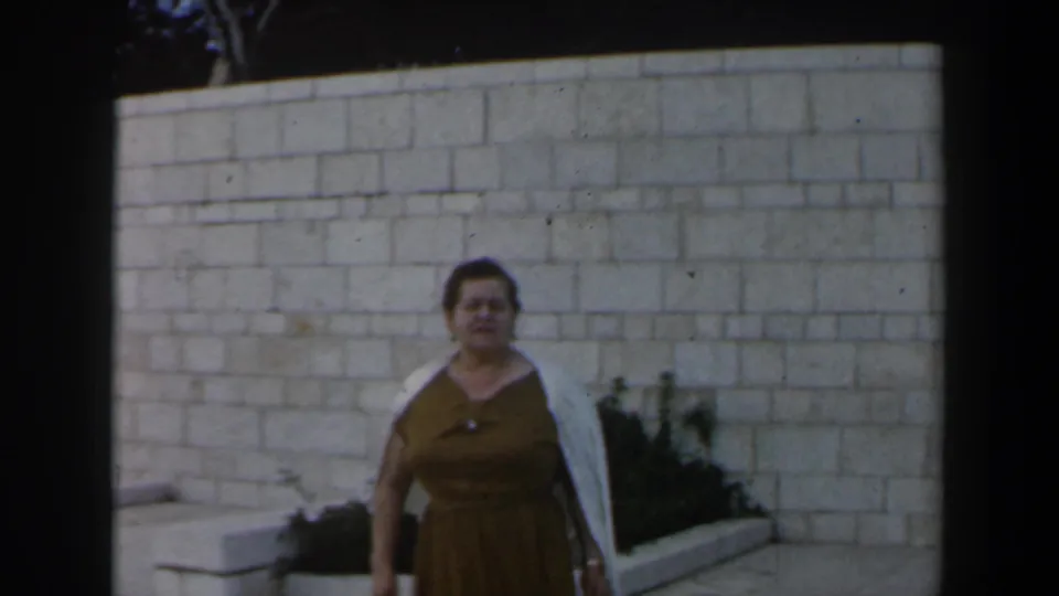 1960s Israel home-movie frame with an adult woman standing in front of the stone-block wall.