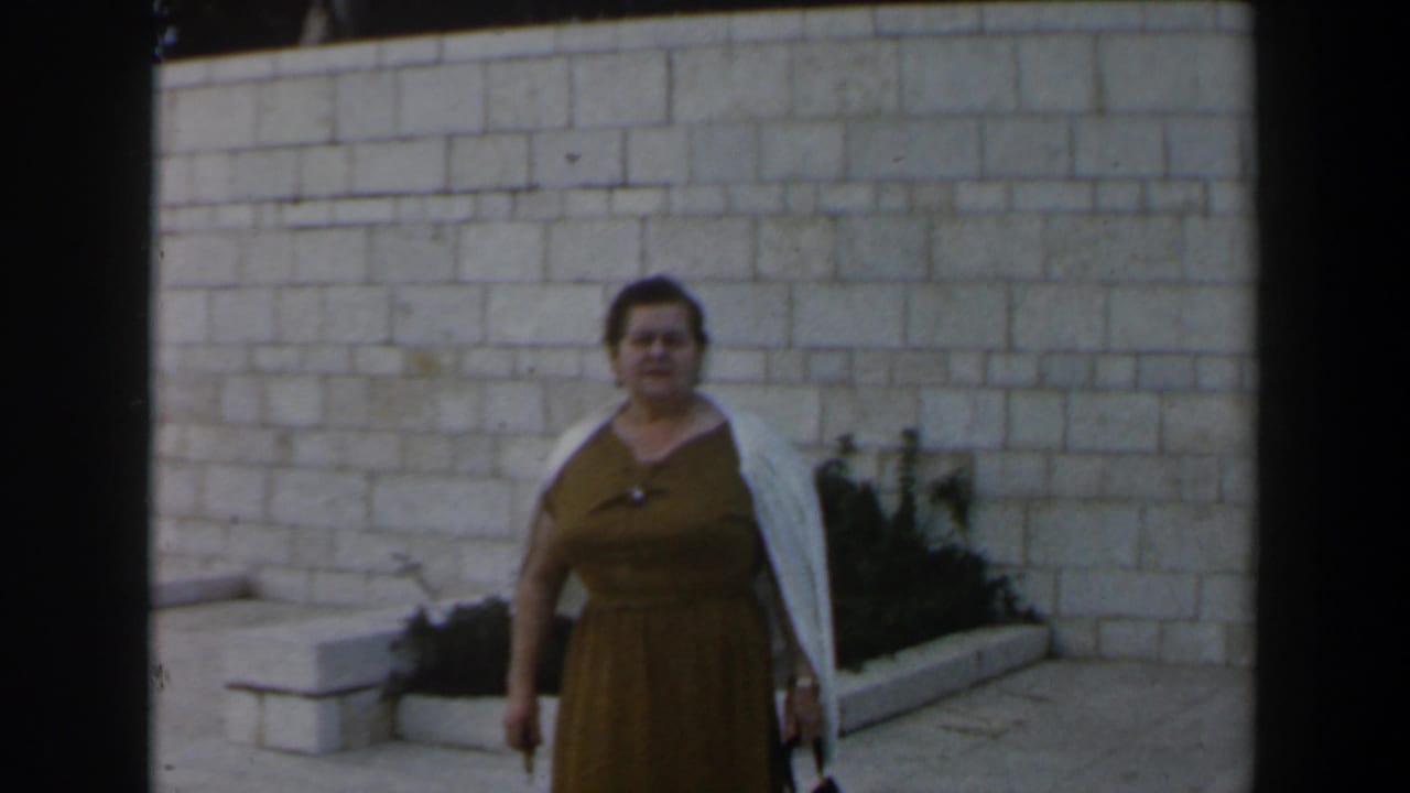 Adult woman in a brown dress and white shawl poses outdoors in Israel before a pale stone wall and raised garden bed.