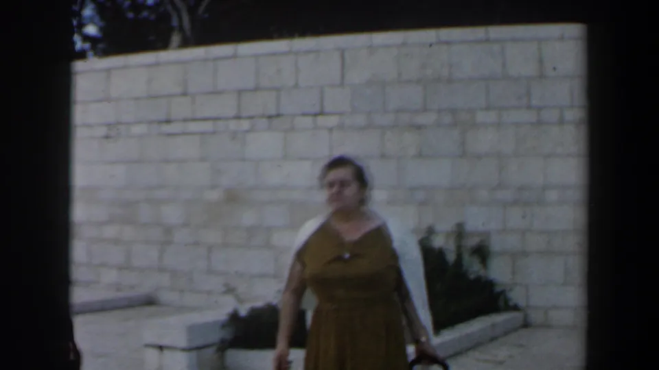 The woman in a brown dress appears slightly farther right, moving or posing near the stone wall in 1960 Israel footage.