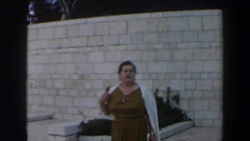 In the 1960 Israel home-movie frame, the woman faces the camera with a white shawl over her shoulder beside a planted bed.