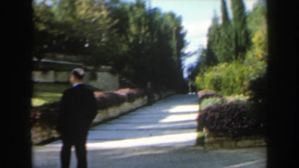 1960 Israel 8mm frame showing a man in a dark suit standing on a landscaped garden path among shrubs and tall trees.