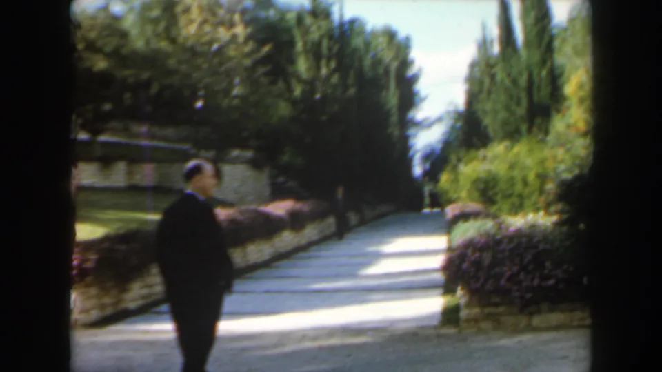 1960 Israel 8mm frame showing a man in a dark suit near a pale garden path with hedges, stone edging, and tall trees.