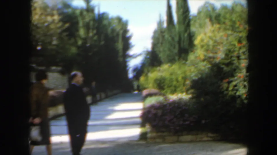 1960 Israel 8mm frame with two adults on a landscaped walkway bordered by hedges, low stonework, and cypress-like trees.