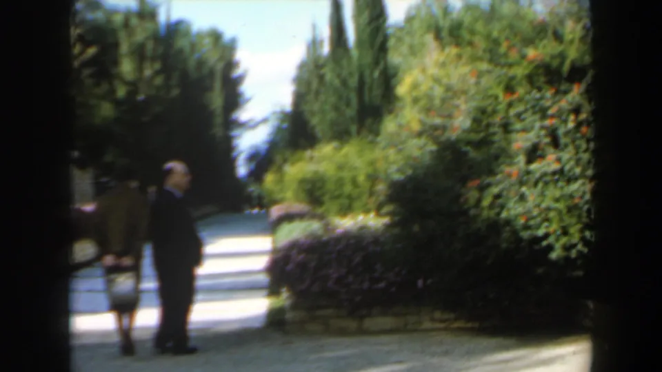 1960 Israel 8mm frame showing a woman in brown clothing and a suited man near a hedge-lined walkway in bright daylight.
