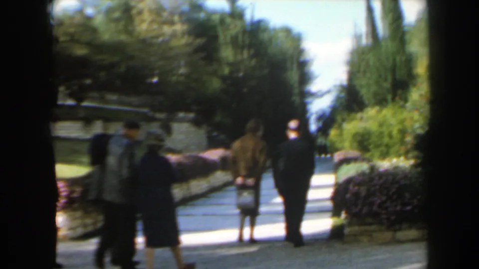 1960 Israel 8mm frame with a small group of adults walking away on a garden path framed by hedges, stonework, and tall trees.