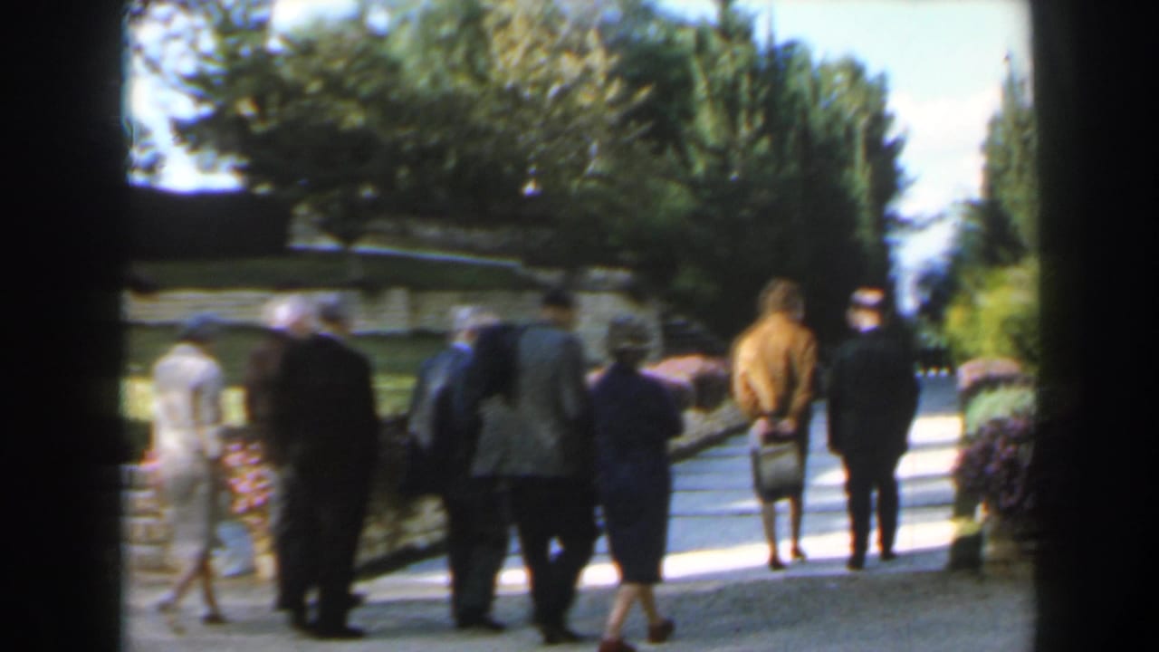 In Israel, 1960, adults in period clothing walk along a paved park path between flowerbeds and low stone retaining walls.