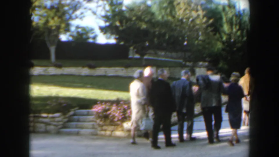 Adults in suits and dresses move through a landscaped Israeli garden, with flowers, trees, and stone edging visible around the path.