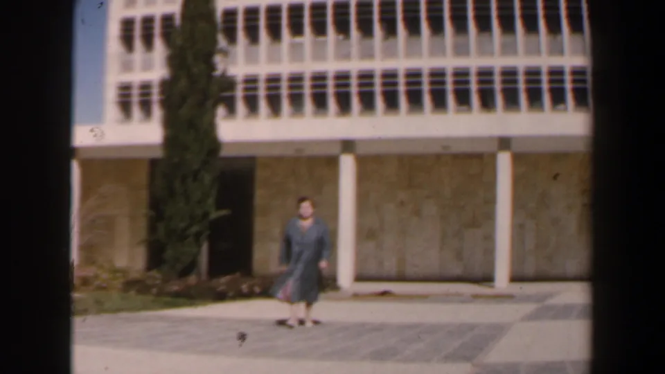 In 1960 Israel footage, a woman in a blue coat appears in front of a modernist exterior with concrete columns, window grid, and cypress tree.