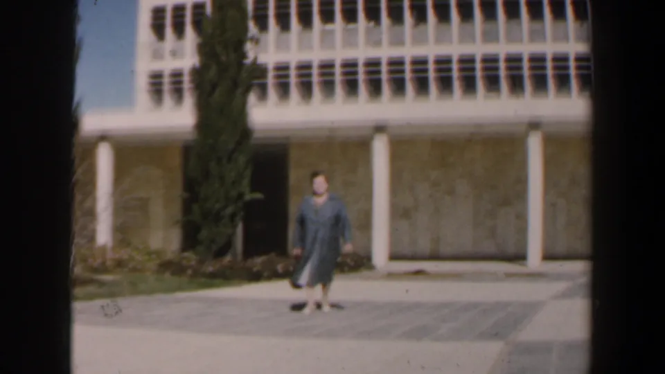 A woman in a blue coat walks toward the camera across a paved courtyard in 1960 Israel, with columns and gridded windows behind her.