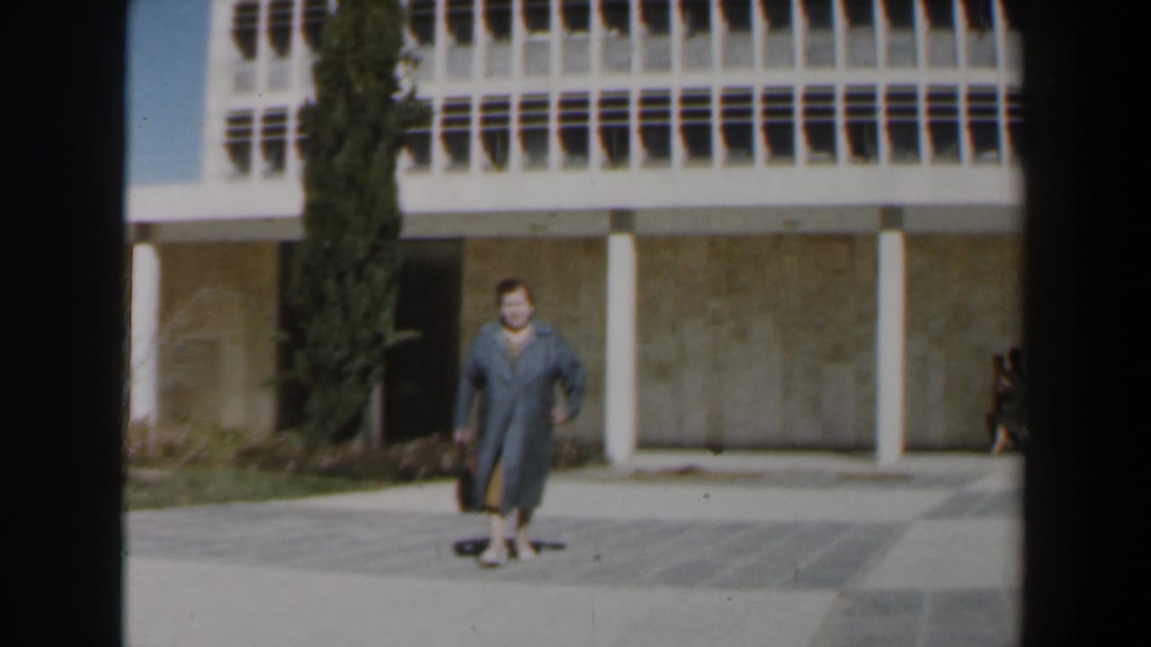 1960 Israel 8mm frame showing a grey-dressed woman with a black bag walking before a white-columned modernist facade and cypress tree.