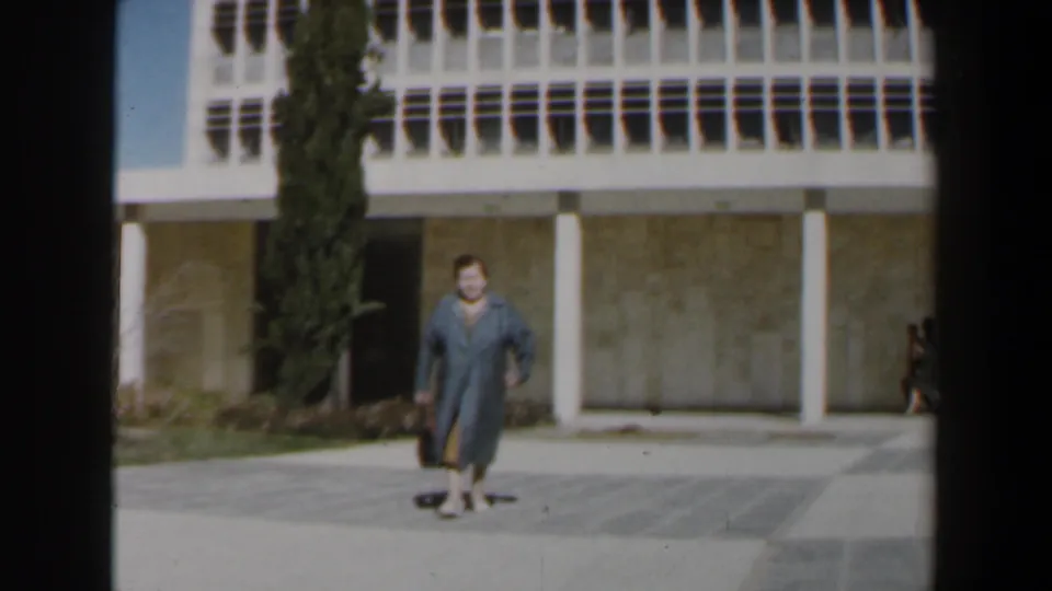 1960 Israel 8mm frame showing a grey-dressed woman with a black bag walking before a white-columned modernist facade and cypress tree.