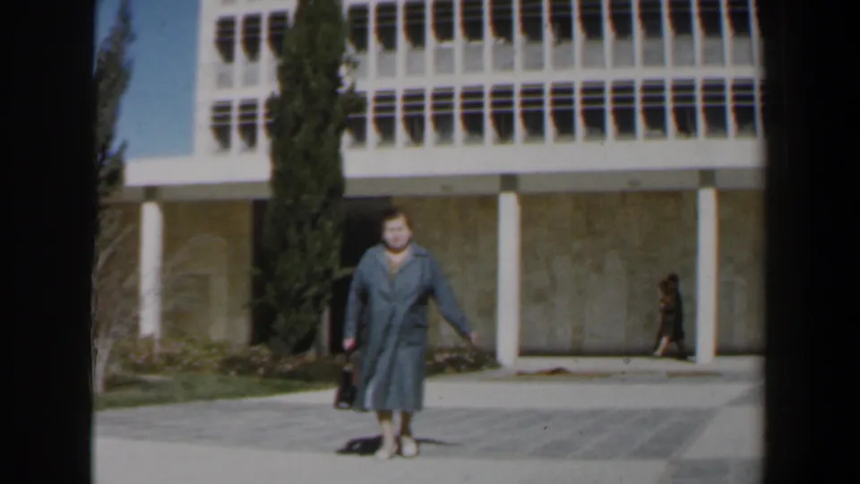 1960 Israel home-movie frame with a woman in a grey dress near the camera, holding a black bag in front of a modernist facade.