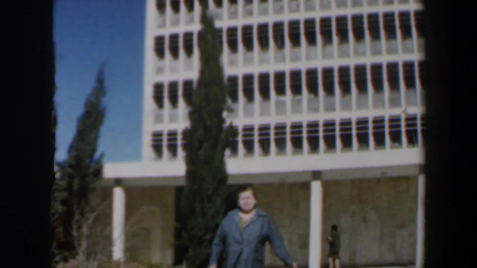 1960 Israel 8mm frame showing the woman nearer the camera, with the modernist building, cypress tree, and a background figure visible.