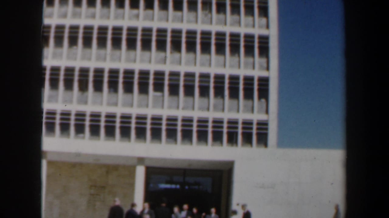 Adults stand near the entrance of a large modern building with gridded windows.