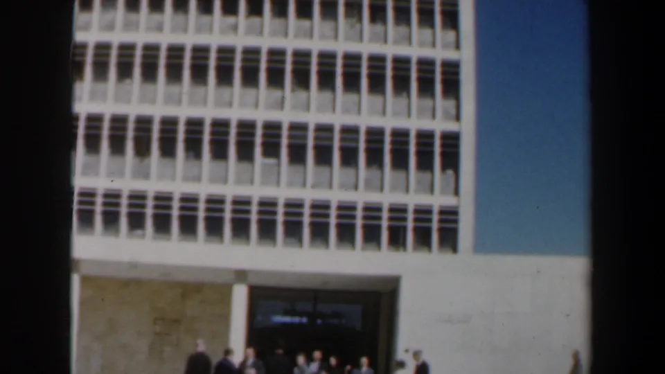 1960s Israel footage shows adults in formal clothing waiting outside a modern concrete building with a recessed entrance and gridded upper windows.