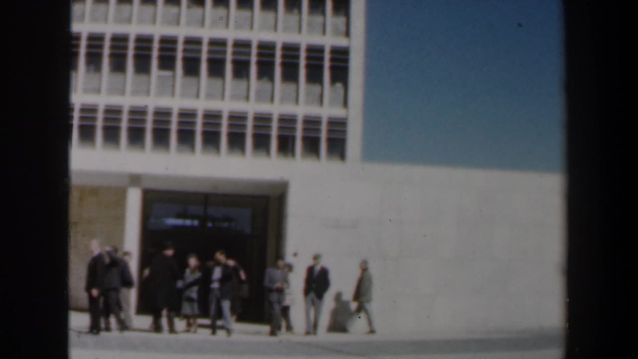 Several adults wait and walk near a large building entrance in daylight.