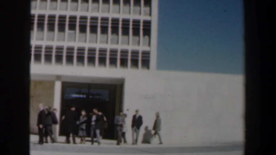 Adults in suits and dresses stand, talk, and pass near the entrance of a large modernist building in Israel, with strong sun on the facade.