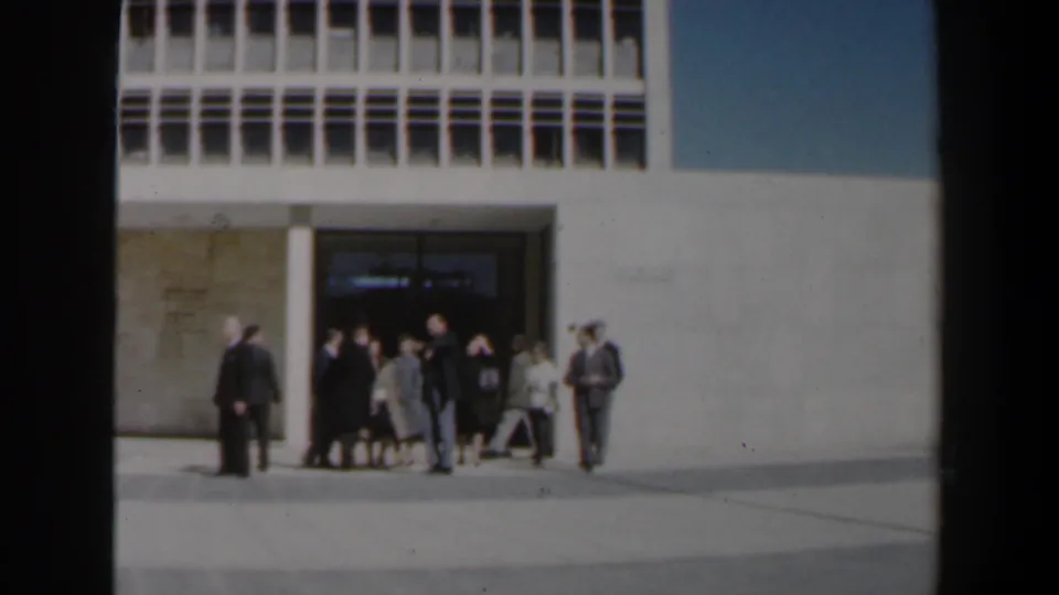 A small group of adults appears to converse outside a large Israeli building, with formal dress, bags, and the entrance visible behind them.