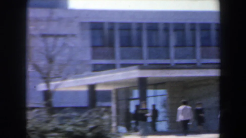 1960 Israel frame showing several pedestrians near a covered entrance, glass doors and a modern concrete facade.