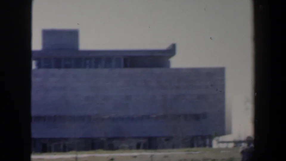 1960 Israel home-movie frame showing a low concrete building with a flat roof, raised roof structure, narrow lower windows, and open foreground ground.