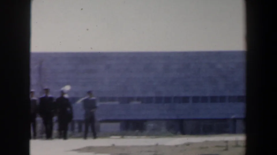 1960 Israel frame with several distant men near a modern concrete building, long horizontal windows, open ground, and soft 8mm blur.