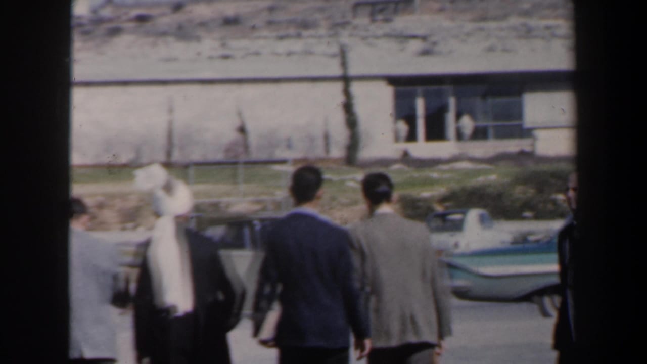 Men walk away from the camera past a low building and parked car.