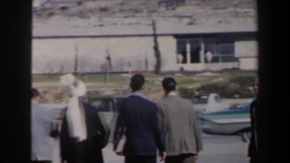 Men in suits and a man with a white head covering walk along a street in Israel around 1960.