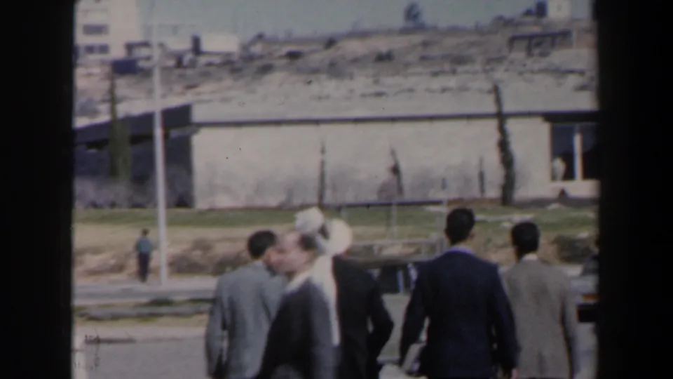 Handheld 8mm view of men walking in a sunlit Israeli street scene around 1960, with one passerby blurred in front.