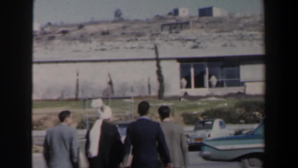 Men in suits and traditional-looking head covering walk past a parked sedan in Israel around 1960.