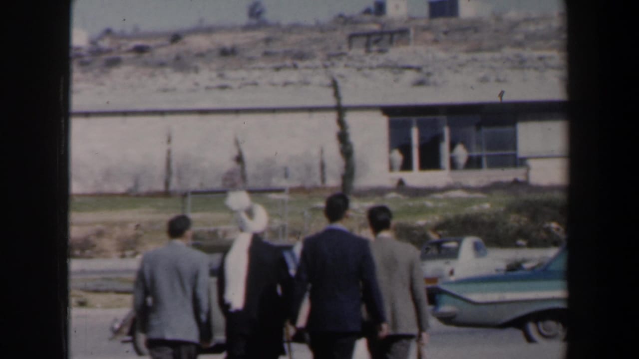 Men in suits walk outdoors near a modern building and a parked teal-and-white car.
