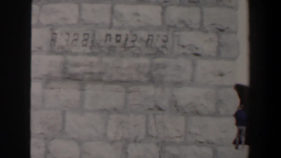 A soft 8mm view of pale masonry with Hebrew writing, with part of a person or dark object entering at the right edge.