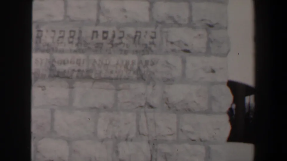 Close 1960 Israel frame of a pale stone wall with Hebrew lettering, partly obscured by dark edge shadow and a partial silhouette.