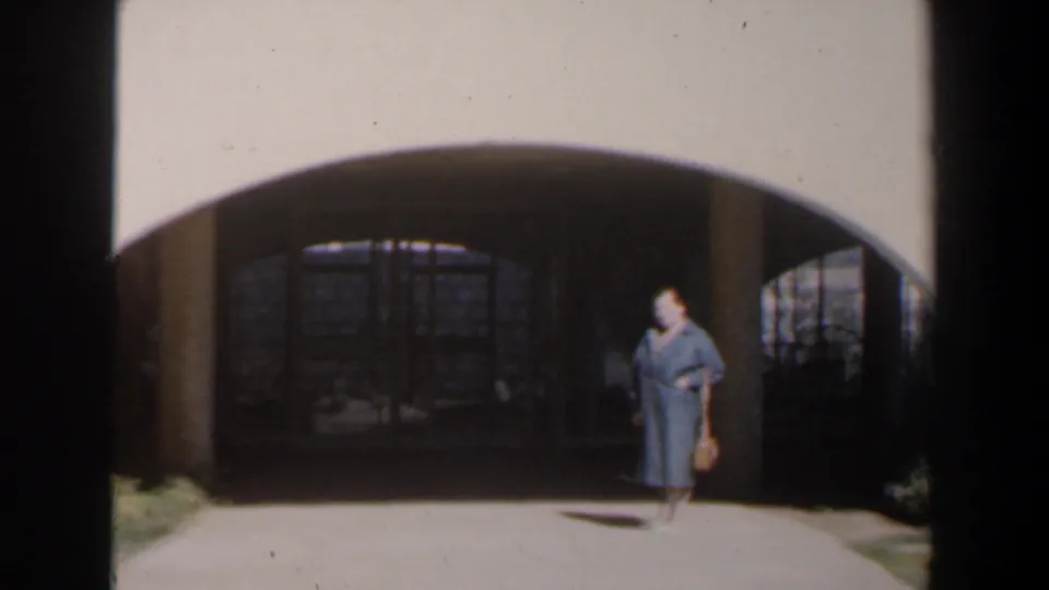 In 1960s Israel, a woman in a long blue dress crosses the sunlit approach to a shadowed stone archway with a basket in hand.