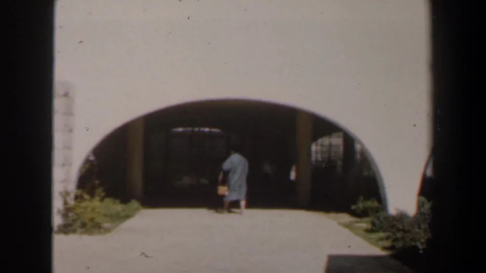 The woman steps into the shadow of the stone archway, with metal gates and courtyard plants visible around the entrance.