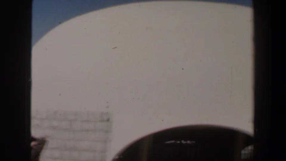 1960 Israel 8mm frame showing a pale arched exterior wall, dark foreground posts, and bright blue sky.