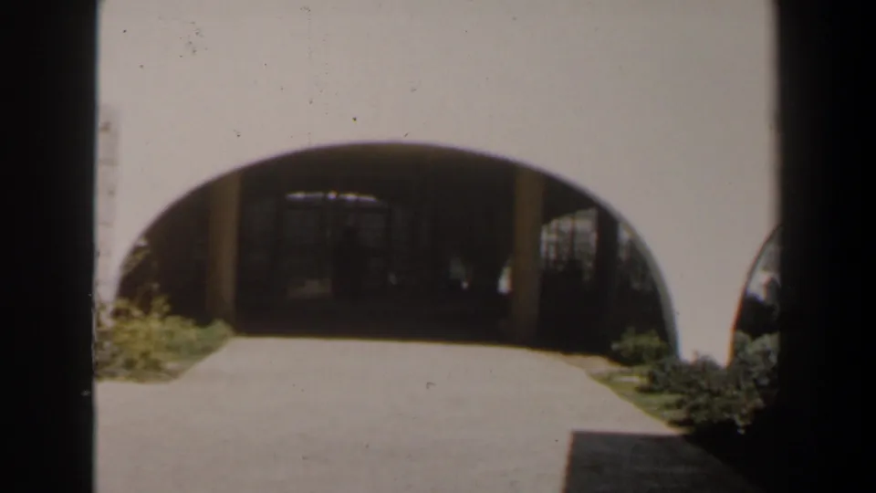 1960 Israel frame showing a sunlit path into a shaded arched entry, with low plants and pale exterior walls.
