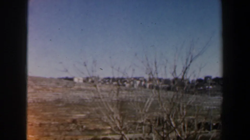 1960 Israel 8mm frame of dry open terrain, leafless foreground trees, scattered distant buildings, and soft faded color.