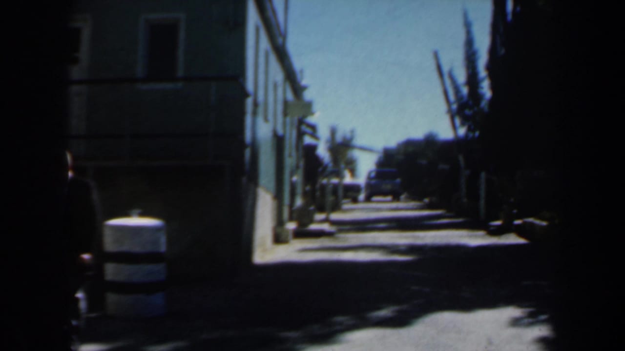 Sunlit narrow alley with pale buildings, trees, striped barrel, and a distant car.
