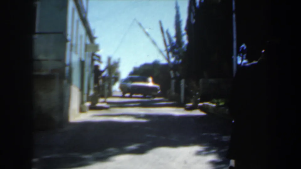 1960 Israel 8mm frame showing a sedan farther along a narrow residential street, with pale walls, trees, and high-contrast sunlight.