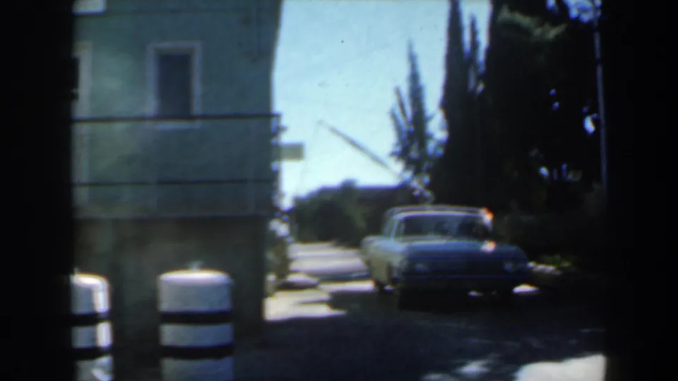 1960 Israel frame with a striped roadside barrel in the foreground and a classic sedan passing through a tree-lined residential alley.