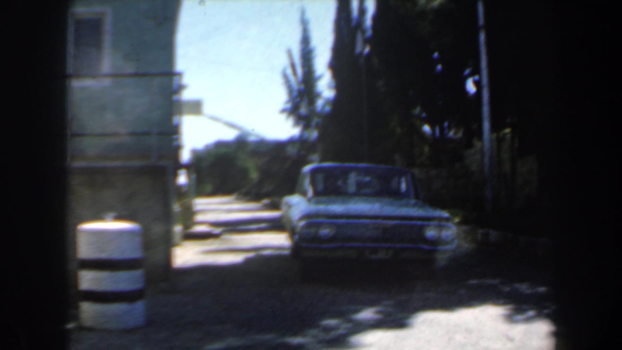 Vintage sedan approaching a narrow gate beside a pale building and striped bollards.