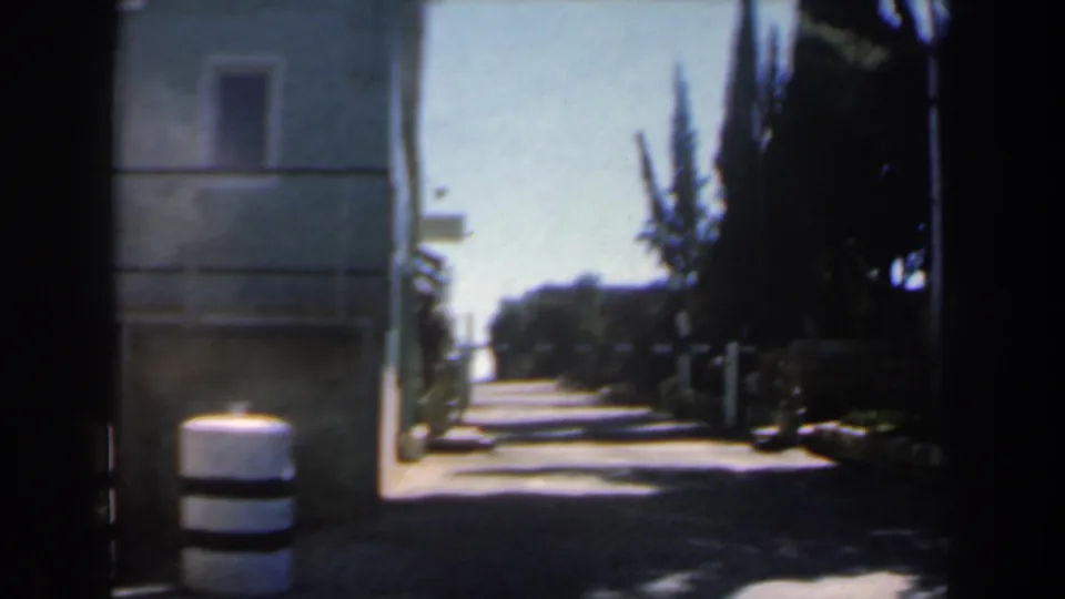 1960s Israel home-movie frame showing the empty gate approach, with a foreground striped bollard, pale building, trees, and strong daylight contrast.