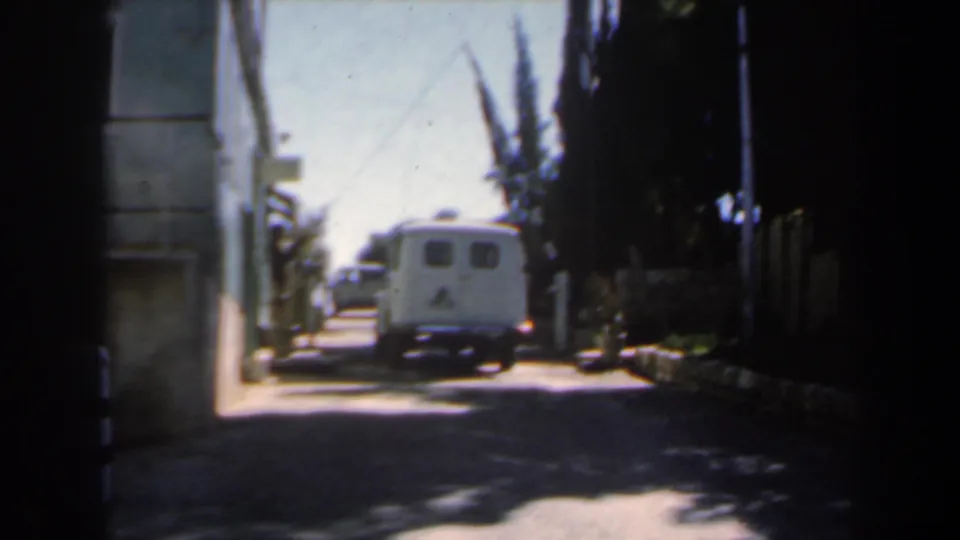 1960s Israel home-movie frame showing a white van moving through the gate area, with bollards, trees, and a pale building along the road.
