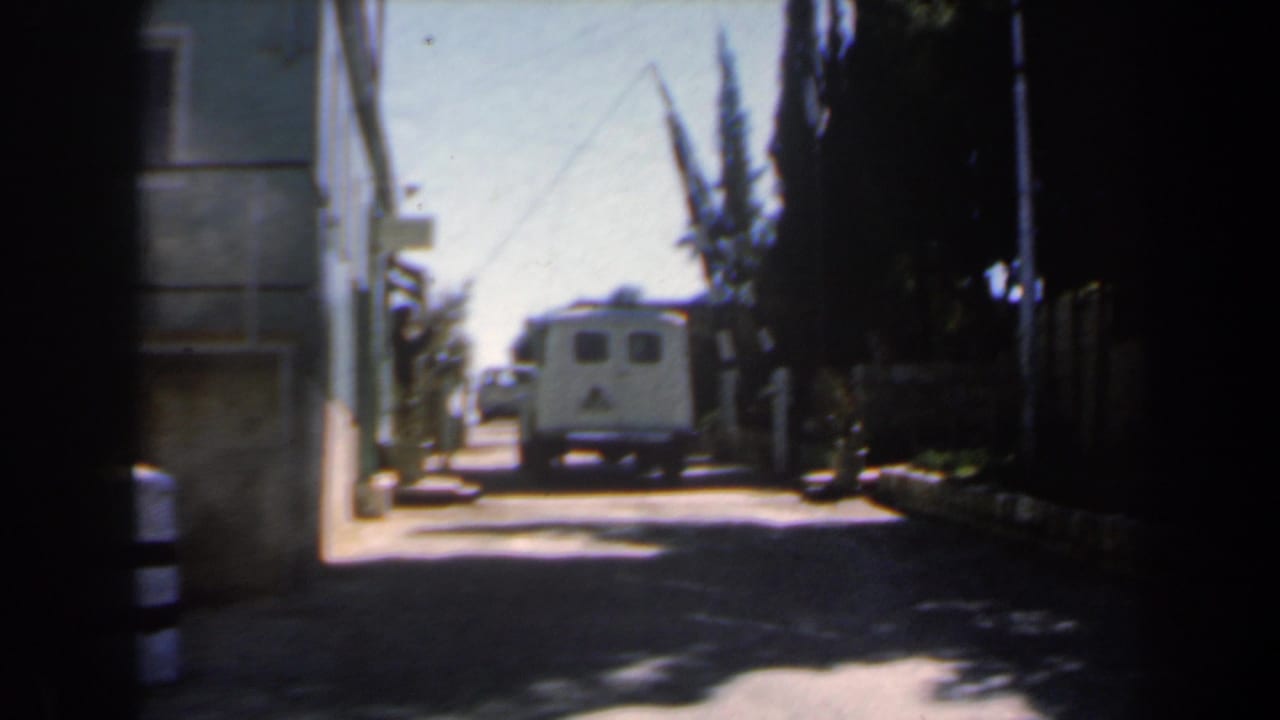 1960s Israel 8mm frame showing a narrow sunlit street from shade, with a white van ahead, building wall at left, and dark trees at right.