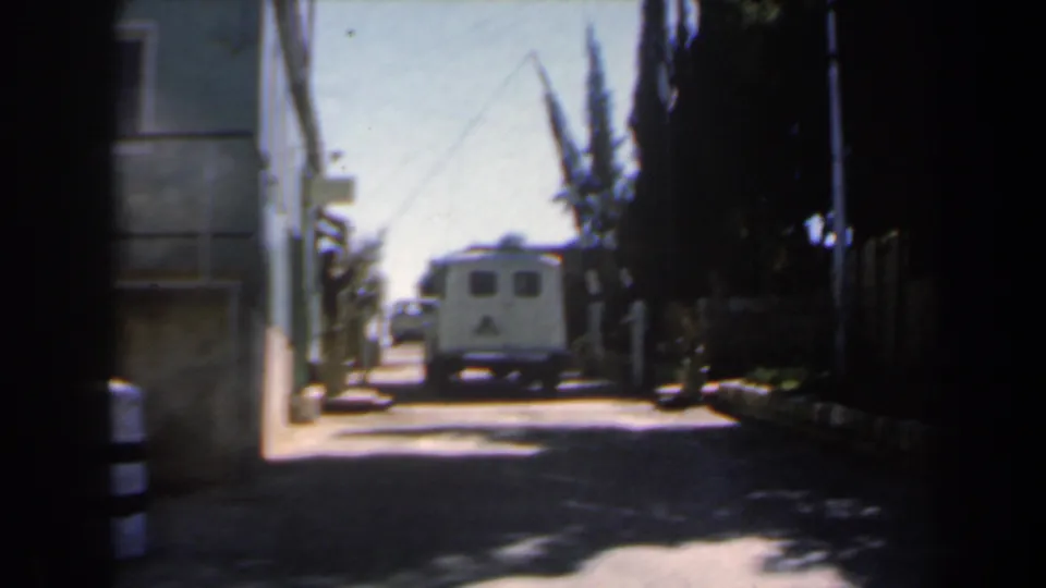 1960s Israel 8mm frame showing a narrow sunlit street from shade, with a white van ahead, building wall at left, and dark trees at right.