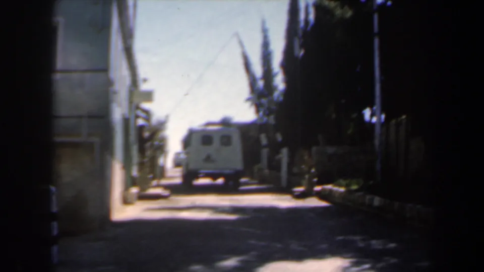 1960s Israel street view with a white van in bright distance, a pale building edge on the left, dark foliage on the right, and high-contrast shade in the foreground.