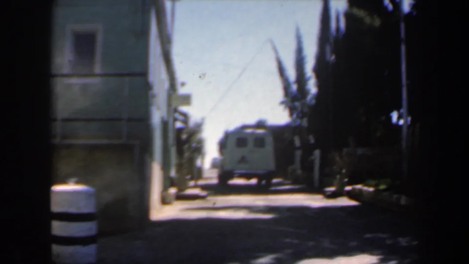 1960s Israel 8mm frame of a narrow street seen from shade, with a distant white van, building frontage at left, tall trees, and a striped post or barrel near the foreground.