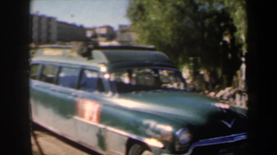 1960s Israel home-movie frame showing a green mid-century car in the foreground, with a narrow street, trees, stone edging, and buildings receding behind it.