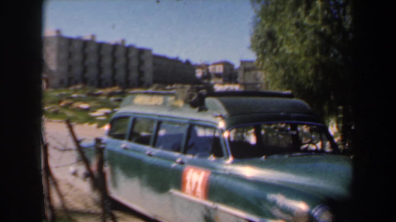 Green station wagon parked beside a fence with apartment blocks in the background.